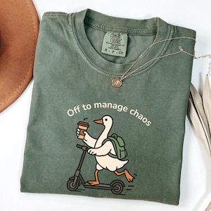 May include: An olive green t-shirt with the text "Off to manage chaos" and a cartoon duck riding a scooter while holding a coffee cup. The shirt also has a Comfort Colors tag and a silver necklace.