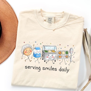 May include: A cream-colored t-shirt featuring a graphic of anthropomorphic lunchroom items including a person in a hairnet, a milk carton, a food warmer, a lunch tray, and a spoon, with the text "serving smiles daily" printed below.