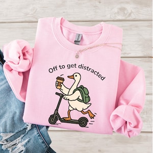 May include: Pale pink sweatshirt featuring the phrase "Off to get distracted." A cartoon goose with a backpack and coffee cup rides a scooter. A gold necklace is draped on the sweatshirt. The design is printed on the front.