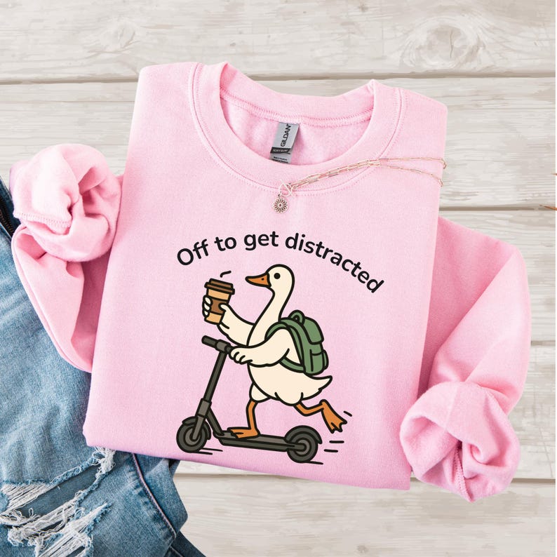 May include: Pale pink sweatshirt featuring the phrase "Off to get distracted." A cartoon goose with a backpack and coffee cup rides a scooter. A gold necklace is draped on the sweatshirt. The design is printed on the front.