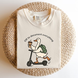 May include: Cream-colored t-shirt featuring a cartoon duck riding a scooter while holding a coffee cup and wearing a backpack. The text "Off to document everything" is printed above the duck. The shirt is folded on a woven surface.
