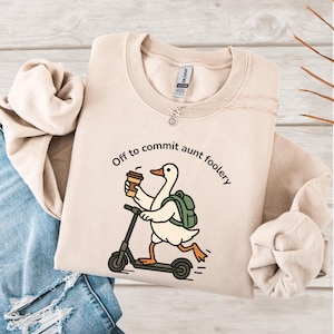 Puede incluir: Sudadera beige con la frase "Off to commit aunt foolery" en texto negro. El diseño presenta un ganso de dibujos animados montado en un scooter mientras sostiene una taza de café. El ganso lleva una mochila verde.