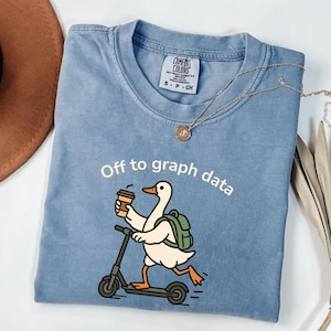 Op de afbeelding: Een lichtblauw Comfort Colors T-shirt met de witte tekst "Off to graph data". De afbeelding toont een cartoon gans op een scooter, met een koffiekopje en een rugzak. Een gouden ketting ligt over het shirt.