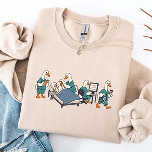 Puede incluir: Una sudadera beige de cuello redondo con un gráfico de cuatro patos blancos vestidos con uniformes médicos de color verde azulado. Un pato sostiene un portapapeles, otro atiende a un paciente en una cama, un tercero está ante un ordenador.