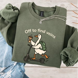 May include: An olive green sweatshirt featuring the text "Off to find veins" and a cartoon duck on a scooter, holding a coffee cup and wearing a backpack. A silver necklace is visible. The sweatshirt is folded.