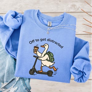 May include: Pale blue sweatshirt featuring the text "Off to get distracted" and a cartoon goose on a scooter, holding a coffee cup. The goose has a backpack. A silver necklace is draped on the sweatshirt.