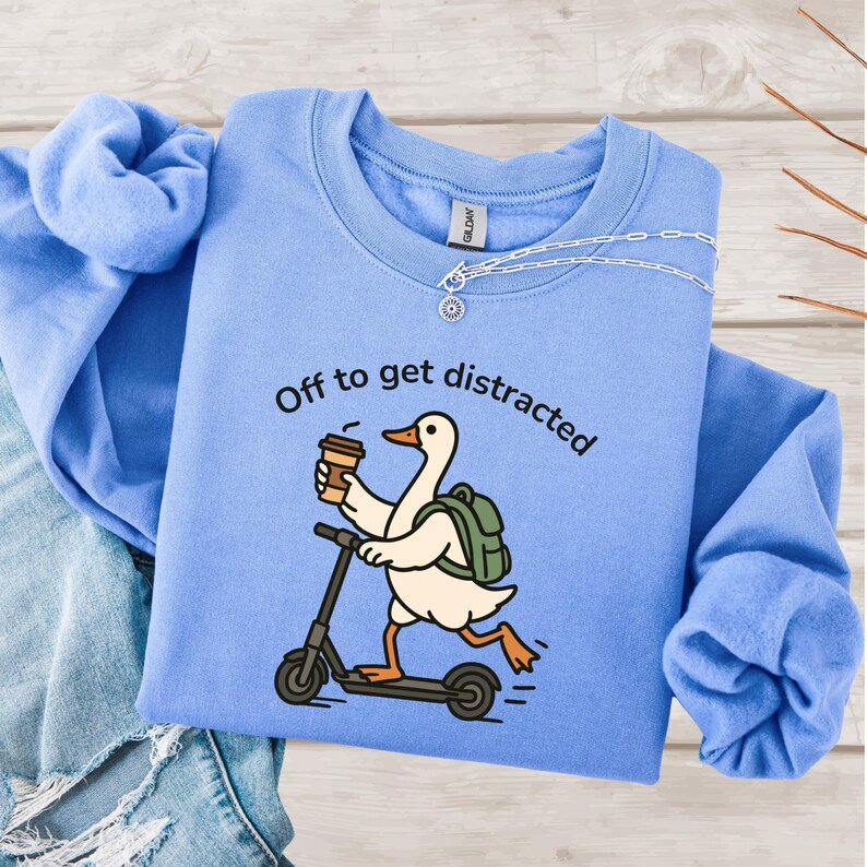 May include: Pale blue sweatshirt featuring the text "Off to get distracted" and a cartoon goose on a scooter, holding a coffee cup. The goose has a backpack. A silver necklace is draped on the sweatshirt.