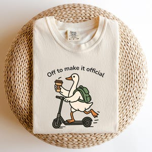 May include: A cream-coloured t-shirt with a cartoon duck on a scooter, holding coffee and wearing a backpack. The text "Off to make it official" is above the duck. The shirt is displayed on a woven, circular surface.