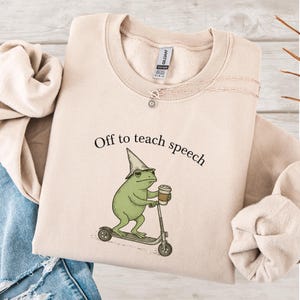 以下が含まれることがあります： 「Off to teach speech」の文字が入ったベージュ色のスウェットシャツ。魔法使いの帽子をかぶり、コーヒーカップを持ち、スクーターに乗った緑色のカエルのグラフィック。太陽のペンダントが付いたネックレスも見える。