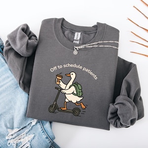 Puede incluir: Una sudadera gris oscuro de cuello redondo con un gráfico de un ganso blanco montado en un patinete, sosteniendo un café y llevando una mochila verde. El texto sobre el ganso dice "Off to schedule patients" en una tipografía blanca.