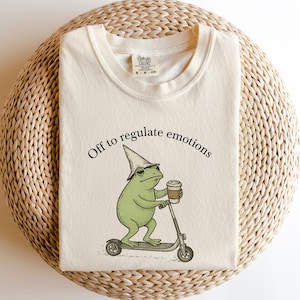 May include: A cream-coloured t-shirt featuring a green frog in a wizard hat, riding a scooter and holding a coffee cup. The text "Off to regulate emotions" is arched above the frog illustration.