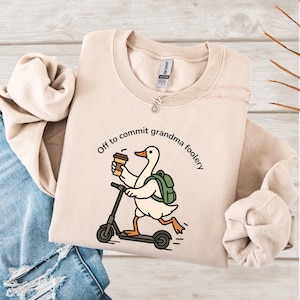 May include: Beige sweatshirt with the phrase "Off to commit grandma foolery." The graphic features a cartoon goose riding a scooter while holding a coffee cup and wearing a backpack. The sweatshirt is on a light wood surface.