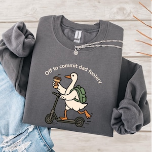 Op de afbeelding: Donkergrijze sweatshirt met de tekst "Off to commit dad foolery" en een cartoon gans op een scooter met een koffiekopje en een groene rugzak. Een zilveren ketting is ook zichtbaar.