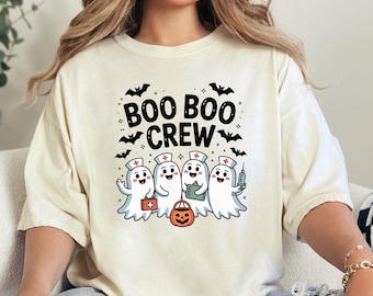 May include: Cream-colored t-shirt featuring the text "BOO BOO CREW" with bats. Below, four cartoon ghost characters dressed as nurses are holding medical supplies and a jack-o'-lantern. A fun Halloween-themed design.