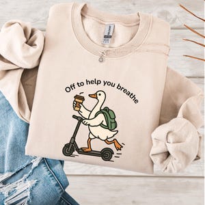 May include: Beige sweatshirt with the text "Off to help you breathe" and a cartoon goose riding a scooter while holding a coffee cup. The goose wears a green backpack. A silver necklace is visible.