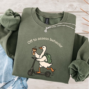 May include: An olive green sweatshirt with the phrase "Off to assess behavior" printed on the front. The graphic features a cartoon duck riding a scooter while holding a coffee cup and wearing a backpack. A silver necklace is also visible.