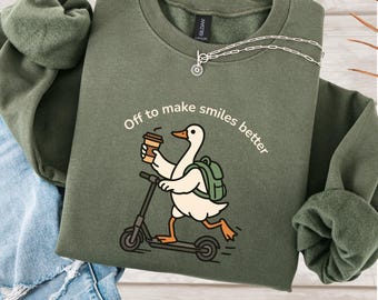 May include: An olive green sweatshirt featuring the phrase "Off to make smiles better." The design depicts a cartoon goose on a scooter, carrying a coffee cup and a backpack. A silver necklace is also visible.