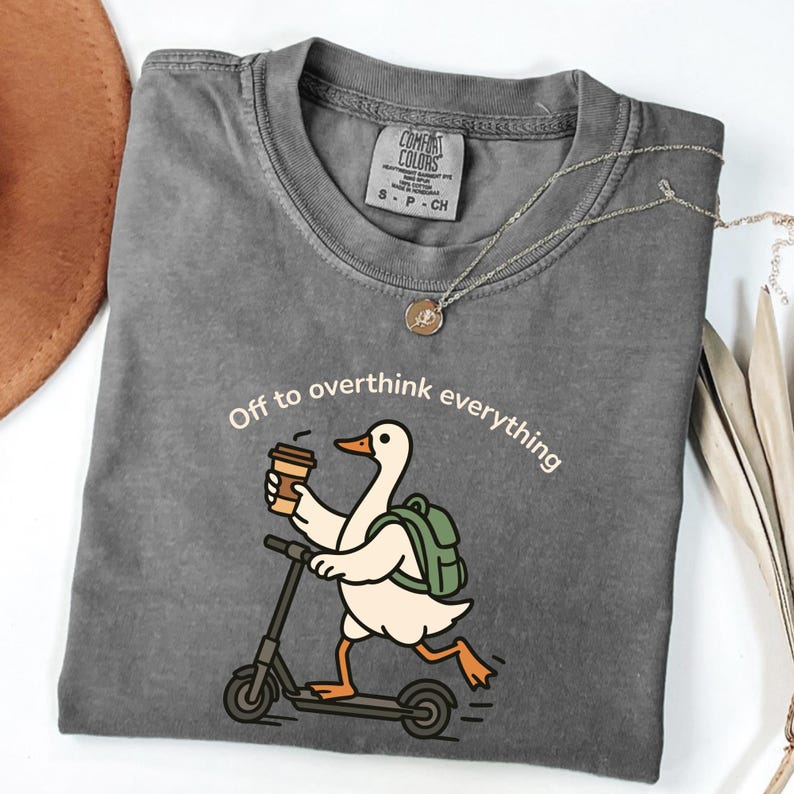 May include: Gray Comfort Colors t-shirt with the phrase "Off to overthink everything." The shirt features a cartoon duck riding a scooter while holding a coffee cup and wearing a backpack. A silver necklace with a pendant is also visible.