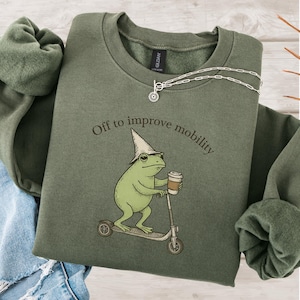 May include: An olive green sweatshirt with the text "Off to improve mobility" above a cartoon frog riding a scooter and holding a coffee cup. A silver necklace is draped across the top of the sweatshirt.
