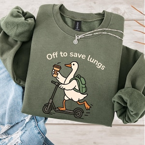 May include: Olive green sweatshirt with the text "Off to save lungs" and a cartoon goose riding a scooter while holding a coffee cup. The goose has a backpack. A silver necklace is on the sweatshirt.