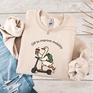 May include: Beige sweatshirt with the phrase "Off to improve mobility" and a cartoon goose riding a scooter while holding a coffee cup. The goose wears a backpack. The sweatshirt is laid on a wooden surface next to a pair of blue jeans.