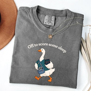 May include: A charcoal grey short-sleeve t-shirt featuring a graphic of a white goose wearing a teal scrub top and a blue shoulder bag. The goose holds a tablet displaying a heart rate monitor. The text reads, Off to score some sleep.