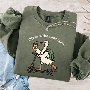 May include: Olive green sweatshirt with the text "Off to write case notes." The graphic features a cartoon goose riding a scooter while holding a coffee cup and wearing a backpack. A silver necklace is also visible.