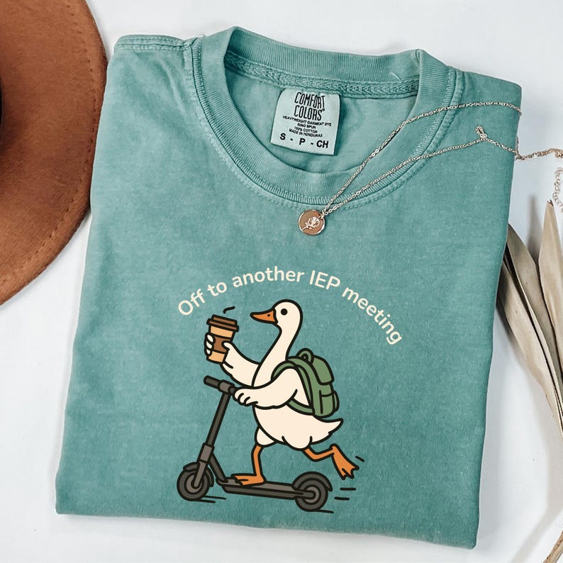 May include: A teal Comfort Colors t-shirt with the text "Off to another IEP meeting." The graphic shows a cartoon duck riding a scooter while holding a coffee cup. A silver necklace with a pendant is also visible.