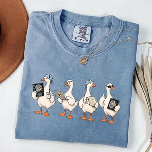 May include: A light blue short-sleeve t-shirt featuring a graphic of four white ducks dressed as medical professionals. The ducks are holding items including an X-ray, a laptop with a radiation symbol, and a mug labeled BEAM IT.