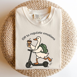 May include: Cream-colored t-shirt with a graphic of a goose riding a scooter, holding a coffee cup, and wearing a backpack. The text "Off to regulate emotions" is above the goose. The shirt is folded on a woven surface.