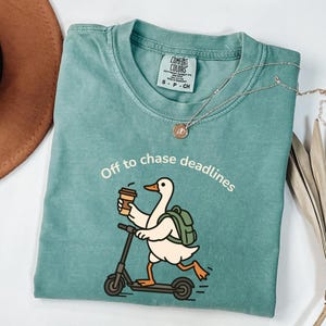 Könnte beinhalten: Ein salbeigrünes T-Shirt mit Rundhalsausschnitt und einer Grafik einer weißen Gans mit grünem Rucksack, die auf einem schwarzen Roller fährt und einen Kaffeebecher hält. Über der Gans steht der Text "Off to chase deadlines" in weißen Buchstaben.