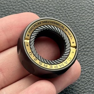 May include: A black and gold EDC Play Mechanical Wheel, a small, cylindrical device. The wheel has the text "EDC PLAY MECHANICAL WHEEL" and "CAMPERS 2.0" in gold lettering. The wheel has a textured, ridged center.