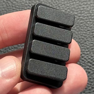 以下が含まれることがあります： 4つの長方形の隆起したセクションを備えた、小さな黒い長方形のオブジェクト。オブジェクトは、灰色の背景に対して人の手で保持されています。オブジェクトはプラスチックまたは同様の材料で作られているようです。