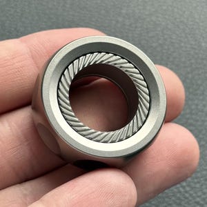 May include: A silver-colored ring with a textured, rope-like design in the center. The ring has a rounded, cylindrical shape with a smooth, matte finish. The ring is held in a person's hand.