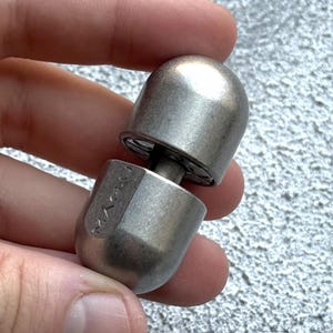 May include: A two-part, silver-coloured metal object with a rounded top and bottom, connected by a central screw. The object is held in a hand against a textured grey background. The top and bottom pieces are both rounded.