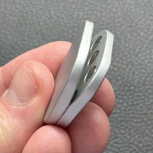 以下が含まれることがあります： 3つの円形の穴と湾曲したデザインの銀色の金属製のオブジェクト。オブジェクトは手で持たれており、その形状と質感が示されています。このアイテムは、小さなツールまたはアクセサリーのようです。