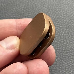 以下が含まれることがあります： 丸みを帯びた細長い形状の、小さな金属製の金色のフィジェットおもちゃ。 おもちゃはヒンジ付きのデザインで、内側に3つの小さな暗い穴が現れます。 物は人の手で持たれています。