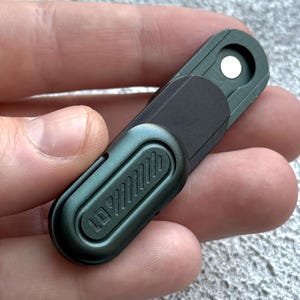 May include: A small, dark green metal tool with a black section and a white circular element. The tool has the text "LD" and a series of angled lines. The tool is held in a hand.