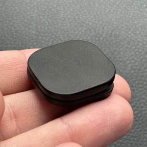 以下が含まれることがあります： 丸みを帯びた角を持つ、小さなマットブラックの正方形の物体。物体は固い素材でできており、わずかに盛り上がったエッジがあります。そのアイテムは人の手で持たれています。