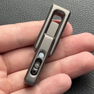 Puede incluir: Un llavero gris metálico con un diseño rectangular. El llavero tiene una sección articulada y una serie de pequeñas incrustaciones circulares. El objeto se sostiene en una mano.