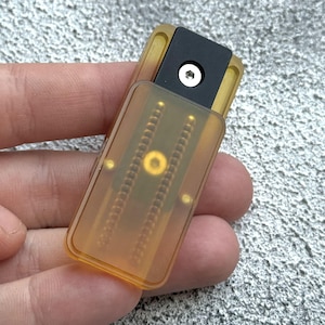 以下が含まれることがあります： 黒い部品と銀色のネジが付いた、小さく長方形の琥珀色の電子デバイス。 デバイスは開いており、内部コンポーネントが明らかになっています。 背景は、テクスチャ加工された明るい灰色の表面です。