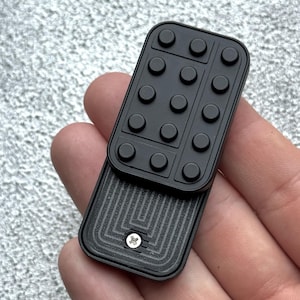 May include: A black fidget toy, rectangular in shape, with a Lego-style design. The top has raised circular studs, and the bottom section features a textured pattern. The toy is held in a hand against a light grey background.