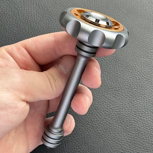 May include: A silver and gold fidget spinner with a long, gray handle. The top of the spinner has a circular design with a gold center and silver outer ring. The handle is cylindrical with black rings near the top and bottom.