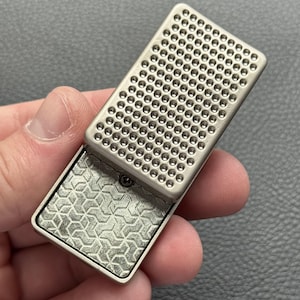 Puede incluir: Un estuche metálico rectangular plateado con tapa abisagrada. La tapa presenta una rejilla de pequeños agujeros espaciados uniformemente. La base abierta del estuche muestra un patrón geométrico. El estuche se sostiene abierto en una mano.