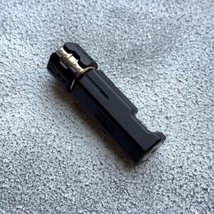 May include: A black and gold metal lighter with the text "ONE TIME" on the side. The lighter has a rectangular body with a rounded top and a gold-colored accent. The lighter is on a textured gray surface.