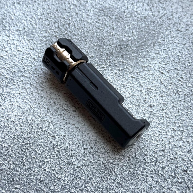 May include: A black and gold metal lighter with the text "ONE TIME" on the side. The lighter has a rectangular body with a rounded top and a gold-colored accent. The lighter is on a textured gray surface.