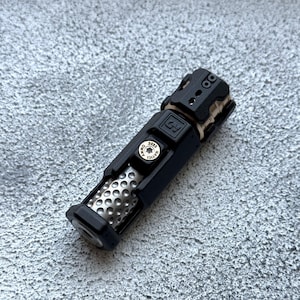 May include: A black and silver metal flashlight with a textured body and a series of small holes. The flashlight has a cylindrical shape with a detailed top and a button that reads "NAVIGI 2023".