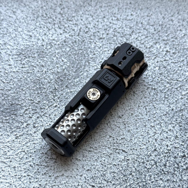 May include: A black and silver metal flashlight with a textured body and a series of small holes. The flashlight has a cylindrical shape with a detailed top and a button that reads "NAVIGI 2023".