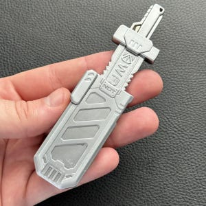 May include: A silver sword-shaped keyring with the text "NOT" and "WAF" etched into the design. The keyring has a rectangular body with a textured surface and a handle with a key ring attachment.
