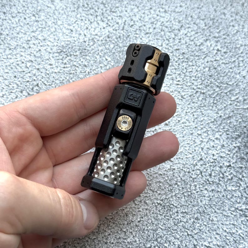 May include: A black and gold metal device with a perforated silver section. The device has a rectangular shape with a hinged top and a circular element with the text "2023". The device is held in a hand.